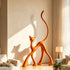 A large, glossy orange sculpture of an abstract cat stands against a neutral wall in a sunlit modern living room. The sculpture features exaggerated, elongated limbs and a high, curved tail, casting a distinct shadow on the wall behind it. Portions of light-colored sofas and a small table lamp are visible in the foreground.