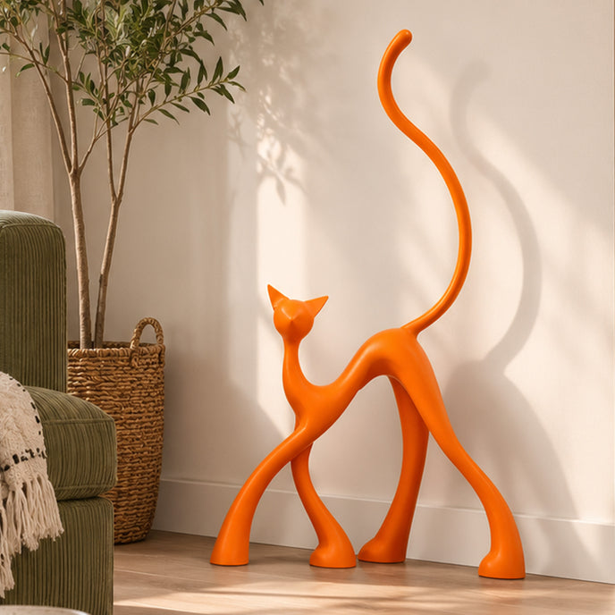 An abstract orange cat-shaped decorative sculpture stands on a light wood floor against a white wall in a styled interior setting with a potted plant, woven basket, and part of a green armchair visible.