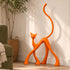 An abstract orange cat-shaped decorative sculpture stands on a light wood floor against a white wall in a styled interior setting with a potted plant, woven basket, and part of a green armchair visible.