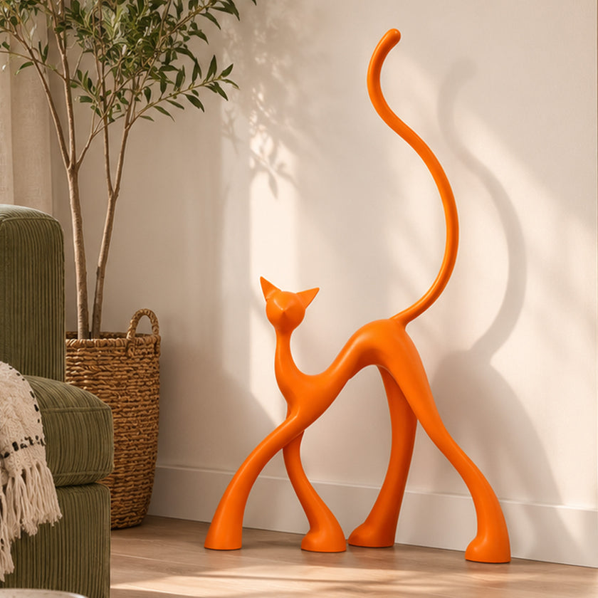An abstract orange cat-shaped decorative sculpture stands on a light wood floor against a white wall in a styled interior setting with a potted plant, woven basket, and part of a green armchair visible.