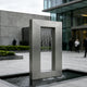 A modern rectangular stainless steel wall fountain with a vertical water cascade, set in a paved outdoor plaza in front of a contemporary glass and stone building.