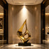 A brilliant hollow golden flame drop stainless steel sculpture displayed in a luxury hallway lobby. This abstract floor art features a fluid, teardrop silhouette with an open center and a highly reflective gold mirror-polished finish. Resting on a sleek black base against a warm, elegant marble interior, the decorative statue radiates modern luxury and sophisticated artistic design.