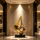 A brilliant hollow golden flame drop stainless steel sculpture displayed in a luxury hallway lobby. This abstract floor art features a fluid, teardrop silhouette with an open center and a highly reflective gold mirror-polished finish. Resting on a sleek black base against a warm, elegant marble interior, the decorative statue radiates modern luxury and sophisticated artistic design.