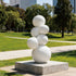A modern outdoor sculpture featuring stacked white spheres on a square concrete base. Displayed on a bright green lawn in a sunny urban park, this minimalist geometric art piece has a smooth, white painted finish. The interconnected round globes create an organic, bubbling effect, offering a striking visual contrast against the natural landscape and background trees.