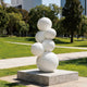 A modern outdoor sculpture featuring stacked white spheres on a square concrete base. Displayed on a bright green lawn in a sunny urban park, this minimalist geometric art piece has a smooth, white painted finish. The interconnected round globes create an organic, bubbling effect, offering a striking visual contrast against the natural landscape and background trees.