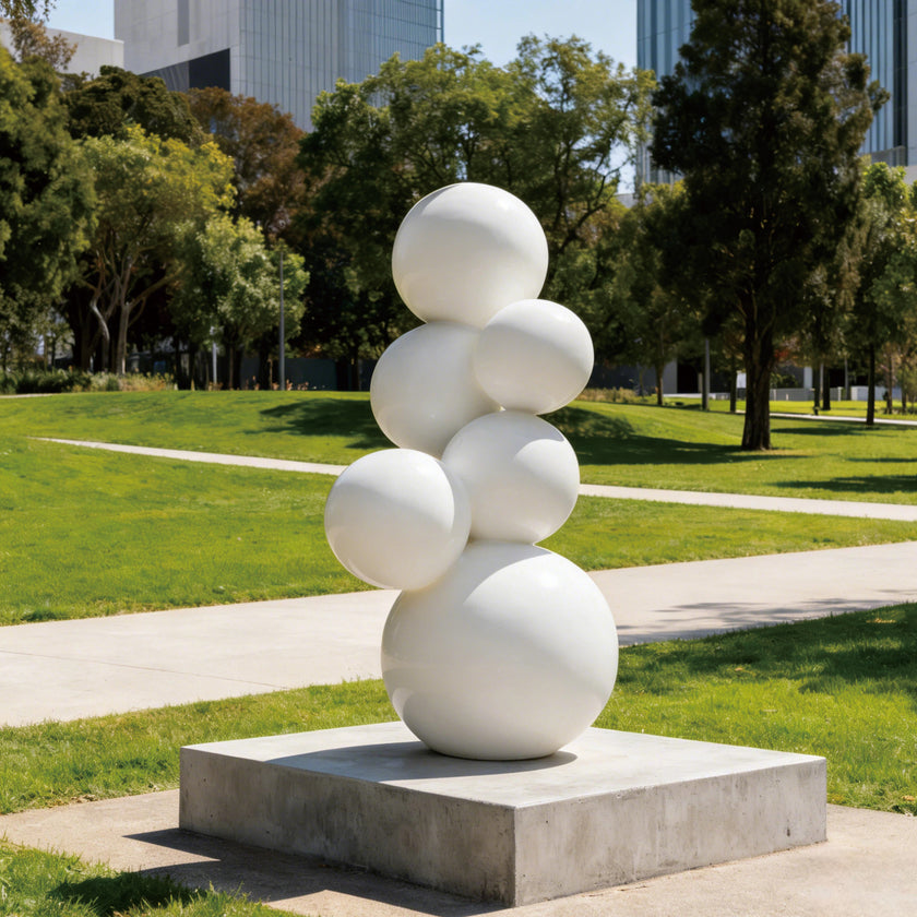 A modern outdoor sculpture featuring stacked white spheres on a square concrete base. Displayed on a bright green lawn in a sunny urban park, this minimalist geometric art piece has a smooth, white painted finish. The interconnected round globes create an organic, bubbling effect, offering a striking visual contrast against the natural landscape and background trees.