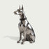 A polished silver metal sculpture of a sitting Doberman dog wearing a black collar with silver studs, set against a solid light gray background.