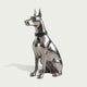 A polished silver metal sculpture of a sitting Doberman dog wearing a black collar with silver studs, set against a solid light gray background.