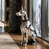 A polished silver metallic dog statue, resembling a Doberman, wearing a black studded collar, displayed on a wooden floor in a modern high-rise office next to large windows with a city view.