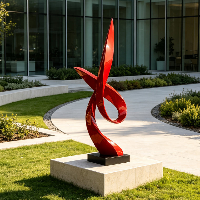 A glossy red abstract spiral sculpture on a black rectangular base, displayed outdoors on a light stone pedestal in a landscaped garden with a modern glass building in the background.