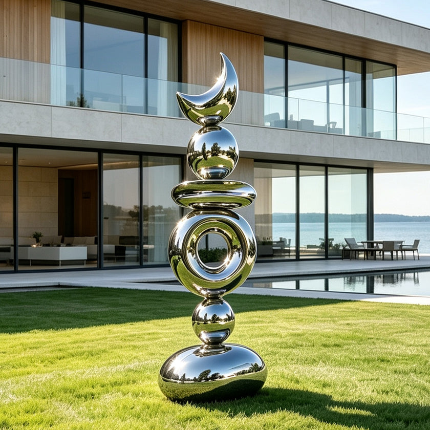 A tall mirror-polished stainless steel sculpture standing on a bright green lawn outside a luxury waterfront villa. This modern abstract art piece features a vertical stack of geometric shapes, including reflective spheres, a hollow ring, and a striking crescent moon at the very top. The shiny silver surface beautifully mirrors the surrounding grass, water, and clear sky, serving as an elegant contemporary outdoor centerpiece.