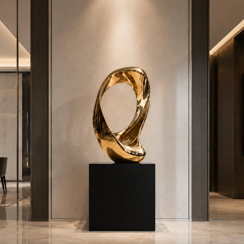 A mirror-polished gold stainless steel sculpture displayed in a luxury foyer. This abstract art piece features an endless, twisted fluid loop with a hollow center, resting on a black square base. The highly reflective golden ring adds a warm, modern elegance to the indoor space.
