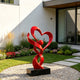 A glossy red stainless steel sculpture in a modern residential courtyard. The abstract design features intertwined ribbons forming a romantic heart shape around a central sphere. Resting on a black square base, this vibrant metal artwork creates a striking focal point among the garden greenery.