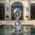 A mirror-polished stainless steel water fountain sculpture in a modern pool. The design features a twisting figure-eight infinity loop with water cascading down its open center. This reflective metal art piece adds a luxurious, dynamic touch to the contemporary estate patio.