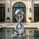 A mirror-polished stainless steel water fountain sculpture in a modern pool. The design features a twisting figure-eight infinity loop with water cascading down its open center. This reflective metal art piece adds a luxurious, dynamic touch to the contemporary estate patio.