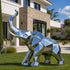 Large modern geometric stainless steel elephant sculpture standing on a green lawn outside a luxury contemporary villa. The elephant features a raised, upward-curving trunk, prominent angular ears, four sturdy legs, and a body constructed from multiple triangular and polygonal facets. Its brilliant mirror-polished silver finish reflects the surrounding greenery, blue sky, and architectural details. The sculpture is displayed in front of a sophisticated residence with natural stone walls, large glass windows
