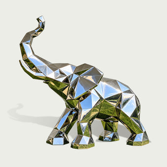 Three-quarter view of a contemporary low-poly stainless steel elephant sculpture featuring a raised trunk, broad geometric ears, and a strong four-legged stance. The entire elephant is formed from precisely defined triangular and polygonal surfaces, creating a distinctive faceted appearance. Its highly reflective mirror-polished silver finish captures blue sky and green landscape reflections across the angular metallic panels. Presented against a clean light background, the sculpture highlights its modern g