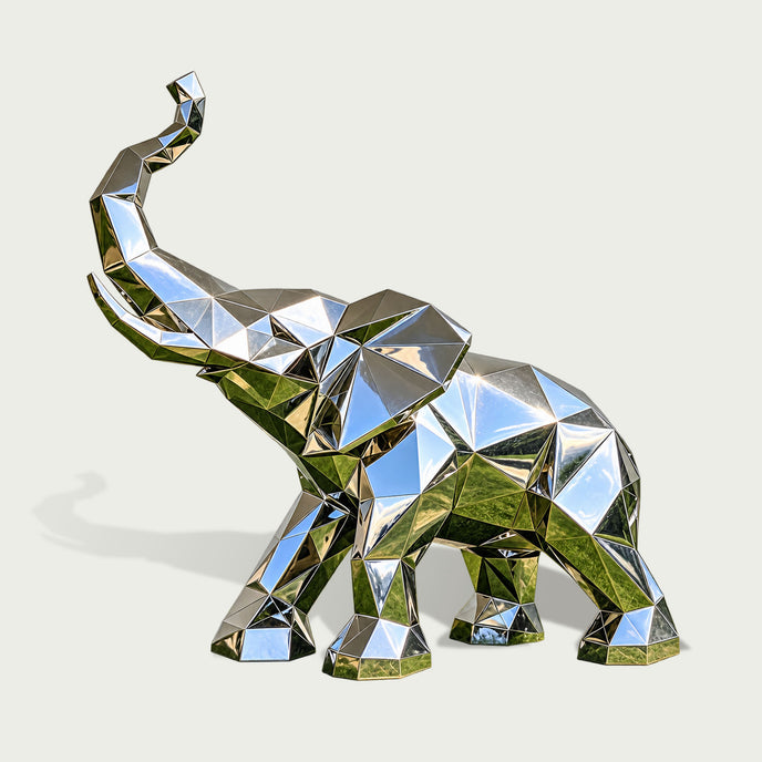 Three-quarter view of a contemporary low-poly stainless steel elephant sculpture featuring a raised trunk, broad geometric ears, and a strong four-legged stance. The entire elephant is formed from precisely defined triangular and polygonal surfaces, creating a distinctive faceted appearance. Its highly reflective mirror-polished silver finish captures blue sky and green landscape reflections across the angular metallic panels. Presented against a clean light background, the sculpture highlights its modern g