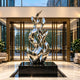 Large modern abstract stainless steel sculpture displayed as a centerpiece in a sophisticated luxury hotel lobby. The artwork features several intertwined, vertically flowing metallic ribbons with elegant leaf-like tips and sweeping organic curves, forming a dramatic upward-reaching composition. Its mirror-polished silver finish reflects the warm interior lighting, glass entrance doors, and surrounding greenery. Positioned on a polished black rectangular pedestal within a recessed floor display area, the sc