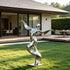 Abstract stainless steel dancer sculpture with outstretched arms, a rounded head and a flowing ribbon-like body, mounted on a square metal base on a garden lawn beside a modern house and wooden terrace.