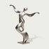 Full view of an abstract stainless steel dancer sculpture with a tilted rounded head, sweeping arms and a twisting ribbon-like torso forming an open loop, finished with brushed silver surfaces and polished accents on a square base against a light background.