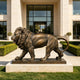 A cast bronze sculpture of a roaring lion stepping on a smooth sphere. This muscular, fierce animal statue is displayed outdoors on a textured stone-like base, serving as a powerful and classic architectural guardian for a luxury estate entrance.